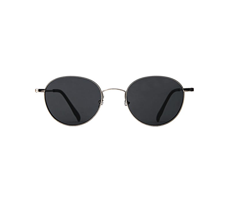 May include: Oval sunglasses featuring dark lenses and a silver metal frame. The design is classic and versatile. The dark lenses offer protection from sunlight. The frame is a thin, silver metal. The sunglasses are suitable for various styles.
