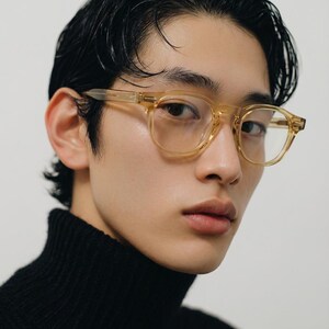 Puede incluir: Gafas transparentes de montura cuadrada con un tinte &aacute;mbar claro. Las gafas est&aacute;n sobre un fondo neutro, y la persona lleva un jersey de cuello alto negro.