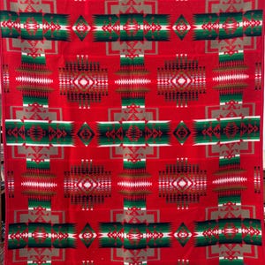 Op de afbeelding: Een rode deken met een geometrisch patroon in groen, wit en grijs. Het ontwerp bevat herhalende kruisvormige vormen en diamantpatronen, waardoor een symmetrisch en visueel opvallend textiel ontstaat.