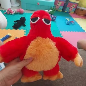 May include: A plush toy in the shape of a red creature with an orange belly, arms, and feet. The toy has large, black-rimmed eyes and is being held by a person. The background includes a play mat and other toys.