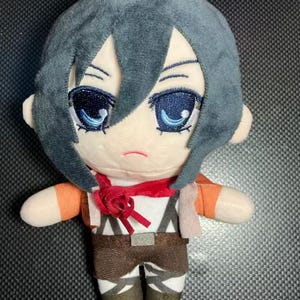 May include: A plush doll of a character with gray hair, blue eyes, and a red scarf. The doll is wearing a white shirt, brown pants, and orange sleeves. The doll has a detailed design and is made of soft fabric.