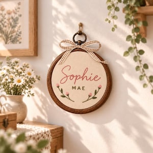 Könnte beinhalten: Ein runder Stickrahmen aus Holz mit cremefarbenem Stoff, auf dem der Name Sophie Mae mit rotem und schwarzem Garn gestickt ist. Das Design zeigt kleine rosa Blumenmotive und eine beige-weiß karierte Schleife am oberen Rand.