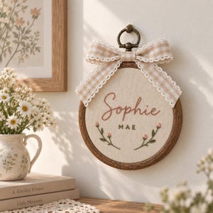 May include: A circular wooden embroidery hoop wall hanging featuring the embroidered name Sophie Mae in pink and dark green thread. The design includes two small floral sprigs and a beige gingham ribbon bow with lace trim attached to the top ring.