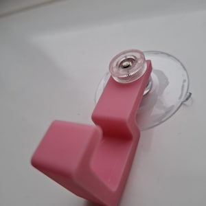 Puede incluir: Un gancho de plástico rosa con una ventosa transparente. El gancho es de un tono rosa claro y tiene una perilla transparente y estriada en la parte superior. La ventosa es redonda y transparente, diseñada para adherirse a una superficie lisa.