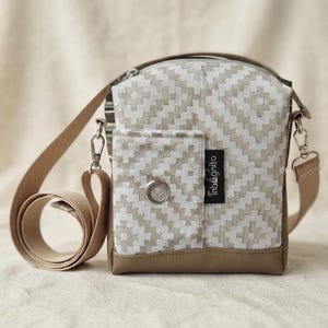Könnte beinhalten: Eine Umhängetasche mit beigefarbenem und weißem geometrischem Muster auf dem Hauptteil und einem einfarbigen hellbraunen Boden. Sie verfügt über eine Vordertasche mit silberner Metallöse, ein schwarzes Etikett mit dem Text inbagnito und einen verstellbaren Riemen.