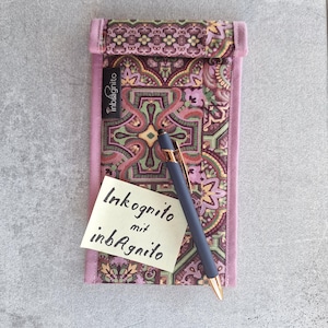 Op de afbeelding: Een rechthoekig stoffen etui met een geometrisch patroon in paars, groen en oranje met een bijpassende paarse rand. Een zwart label toont inbAgnito. Een donkergrijze pen met gouden accenten en een geel briefje met de tekst Inkognito mit inbAgnito.