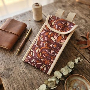 Op de afbeelding: Een rechthoekig stoffen tasje met een herhalend bloemenpatroon in tinten diep bordeaux, gebrand oranje, crème en zachtroze. Het tasje heeft een afgeronde klepsluiting, lichtbeige randen en twee bijpassende stoffen lussen aan de bovenkant.