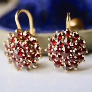 May include: A pair of gold-toned earrings, each featuring a cluster of small, deep red gemstones. The earrings have a floral or starburst design, with a hook closure. The gemstones are closely set, creating a dense, sparkling appearance.