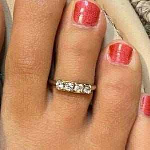 Può includere: Primo piano di un anello d'oro con tre pietre preziose trasparenti disposte in fila. L'anello è su un dito del piede con smalto rosso. L'incarnato è chiaro.