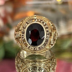 May include: A gold-colored ring with a large, oval, dark red gemstone at the center. The ring is engraved with the words "WEST POINT" and the year "1943". The ring has intricate detailing around the band.