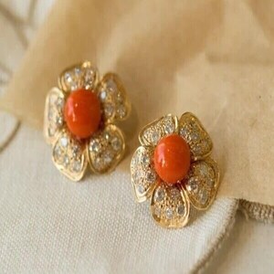 Puede incluir: Un par de pendientes dorados con forma de flor. Cada pendiente presenta una piedra central de color naranja vibrante, rodeada de pétalos incrustados con pequeñas piedras claras y brillantes. Los pendientes se exhiben sobre una superficie color crema.