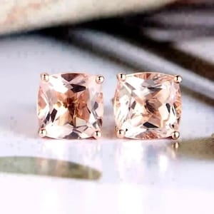 Puede incluir: Un par de pendientes de botón de morganita de corte cuadrado. Las piedras preciosas tienen un tono rosa melocotón y están engastadas en oro rosa. Los pendientes se muestran sobre una superficie blanca, con un fondo borroso.