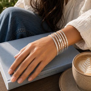 Op de afbeelding: Een set van vijf zilverkleurige armbanden gedragen om de pols. De armbanden hebben verschillende texturen, waaronder gladde oppervlakken, ingewikkelde gegraveerde patronen en een kleine stervormige bedel aan een van de middelste banden.