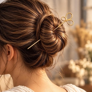 May include: A gold-toned metal hair pin featuring a decorative top with three symmetrical loops arranged in a clover-like shape. The pin is inserted horizontally through a bun of brown hair, securing the style with its slender, straight shaft.