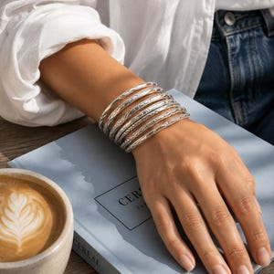 Puede incluir: Un conjunto de seis brazaletes de plata con intrincados patrones grabados. Cada pulsera tiene un acabado metálico pulido con texturas detalladas, llevadas juntas en la muñeca para crear una apariencia superpuesta con tonos plateados.