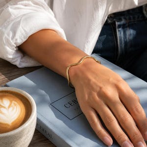May include: A gold-toned metal bangle bracelet featuring a continuous, smooth, undulating wave design. The bracelet has a polished, reflective surface and a rounded profile that encircles the wrist with a fluid, curved shape.