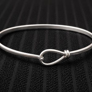 Może przedstawiać: Srebrzysta metalowa bransoletka typu bangle z gładką, okrągłą obręczą, która pośrodku przechodzi w detal w kształcie łezki. Bransoletka ma polerowane, odblaskowe wykończenie oraz jednolitą, solidną konstrukcję na całym obwodzie.