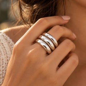 Può includere: Un set di tre anelli impilati color argento con una superficie liscia, lucida e arrotondata. Gli anelli presentano un design a fascia spessa e bombata che si adagia perfettamente l'uno sull'altro, creando un aspetto metallico e riflettente.