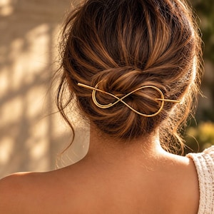 May include: A gold-toned metal hair accessory featuring a smooth, continuous infinity loop design. The accessory consists of a long, straight pin that passes through a looped wire shape, securing a bun of brown hair in place with a polished finish.