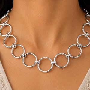 Puede incluir: Un collar de tono plateado que presenta una serie de eslabones circulares uniformes y entrelazados. La cadena está construida con anillos de metal pulido conectados por pequeñas bisagras discretas, creando un patrón circular continuo y simétrico.