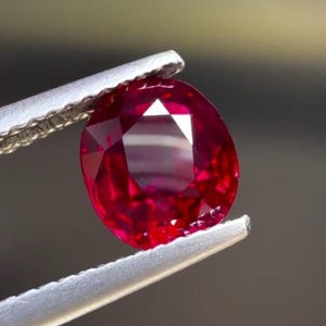 Könnte beinhalten: Ein leuchtend tiefroter Edelstein mit facettiertem Rundschliff. Der Stein wird von einer metallischen Pinzette mit gezackten Kanten gehalten, was seine Transparenz und die durch Lichtreflexionen erzeugten geometrischen Muster hervorhebt.