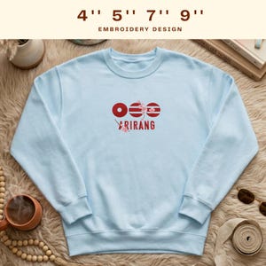 May include: A light blue sweatshirt with the word "ARIRANG" in red and white lettering. Above the word are three red circles and stripes. The text "EMBROIDERY DESIGN" is above the sweatshirt.