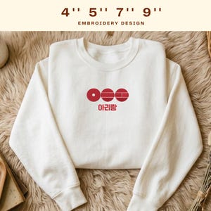 May include: A white sweatshirt with a crew neck featuring a red embroidered design of three circles and the Korean word "Arirang" underneath. The text "4" 5" 7" 9" EMBROIDERY DESIGN" is at the top.