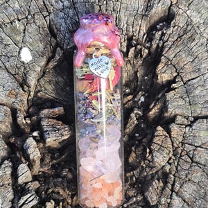 May include: A clear glass bottle filled with layers of pink and purple crystals, dried herbs, and pink wax. A small silver heart charm with the words "made with love" hangs from the bottle. The bottle is sealed with pink wax and glitter.