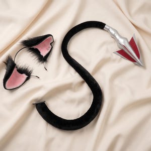 Op de afbeelding: Een zwarte kattenoren-hoofdband met roze binnenoren en een bijpassende zwarte staart. De staart heeft een zilveren en rode pijlvormige punt. De items worden gepresenteerd op een crèmekleurige stof.