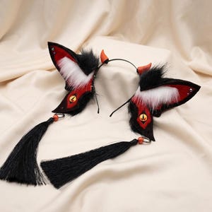 Könnte beinhalten: Ein Paar Cosplay-Fuchsohren in Rot, Schwarz und Weiß mit flauschiger Textur und kleinen orangefarbenen Hörnern. Schwarze Quasten hängen an den Ohren, die an einem schwarzen Stirnband befestigt sind.