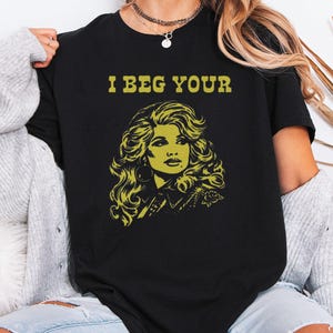 Könnte beinhalten: Ein schwarzes Kurzarm-T-Shirt mit einem gelben Grafikdruck einer Person mit voluminösem Haar und dem Text "I BEG YOUR" über der Abbildung. Das T-Shirt hat einen Rundhalsausschnitt und eine Standardpassform mit einem auffälligen Design.