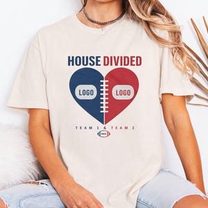 May include: A cream-coloured short-sleeve t-shirt featuring a graphic of a heart split into a navy blue side and a red side. The heart is divided by white football laces. The text reads HOUSE DIVIDED, TEAM 1 & TEAM 2, and includes a small football.
