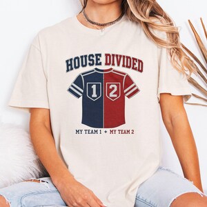 May include: A cream-colored short-sleeve t-shirt featuring a graphic of a split-color jersey in navy blue and red. The text reads HOUSE DIVIDED at the top and MY TEAM 1 + MY TEAM 2 at the bottom, with the numbers 1 and 2 on the jersey halves.