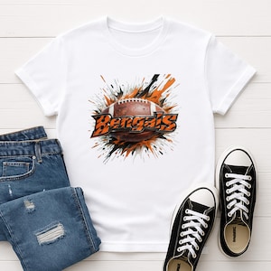 May include: A white short-sleeved t-shirt featuring a graphic print of a brown American football centered over an orange and black paint splatter design. The word Bengals is printed in a bold, stylized orange font with a black outline across the ball.