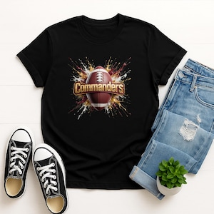 May include: A black short-sleeved t-shirt featuring a central graphic of a brown football with white laces, surrounded by a gold and burgundy paint splatter effect, with the word Commanders printed in gold lettering across the football.