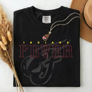 May include: Black t-shirt with the words "INDIANA FEVER" in red and yellow lettering. The shirt features a stylized logo and the Comfort Colors brand tag. A silver necklace with a star pendant is draped on the shirt.