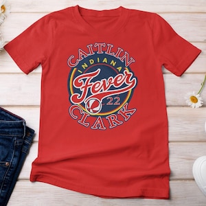 Op de afbeelding: Rood T-shirt met een cirkelvormige afbeelding met de tekst "CAITLIN INDIANA Fever CLARK 22". De afbeelding heeft een marineblauw en geel kleurenschema met een basketbalontwerp. Het shirt is gemaakt van zacht materiaal.