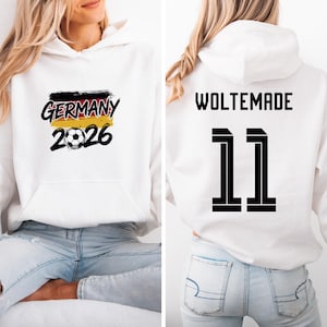 Puede incluir: Sudadera con capucha blanca con un gráfico frontal que presenta "GERMANY 2026" en negro, rojo, amarillo y un balón de fútbol. La parte trasera de la sudadera muestra "WOLTEMADE" sobre el número "11" en negro.