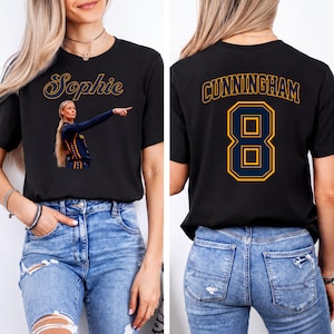 Op de afbeelding: Een zwart t-shirt met korte mouwen met een afbeelding van een basketballer die wijst met de naam Sophie erboven op de voorkant. De achterkant van het shirt toont de naam CUNNINGHAM in een gebogen lettertype boven het grote rugnummer 8.