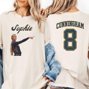 May include: An off-white t-shirt featuring a graphic of a basketball player pointing with the name Sophie above it on the front, and the name CUNNINGHAM with the number 8 in dark blue and gold lettering printed on the back of the garment.