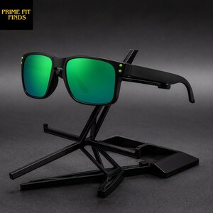 May include: Black sunglasses with green lenses are displayed on a black stand. The sunglasses have a square frame with green lenses. The stand is a geometric design with a small tray at the base. The text "PRIME FIT FINDS" is in the upper left corner.