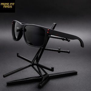 May include: Black sunglasses with dark lenses and red accents on the temples. The sunglasses are displayed on a black metal stand. The text "PRIME FIT FINDS" is in the upper left corner.