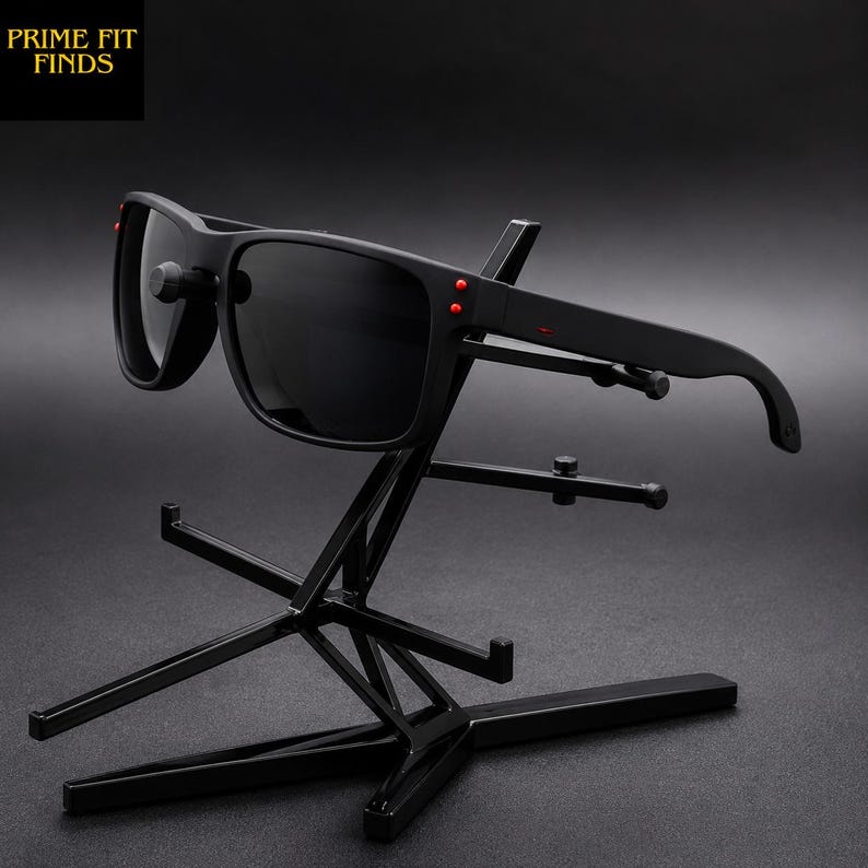 May include: Black sunglasses with dark lenses and red accents on the temples. The sunglasses are displayed on a black metal stand. The text "PRIME FIT FINDS" is in the upper left corner.