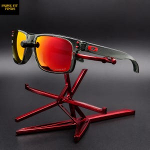 May include: Gray sunglasses with red lenses are displayed on a red metal stand. The sunglasses have a rectangular frame and a small red accent on the temple. The stand has a starburst design.