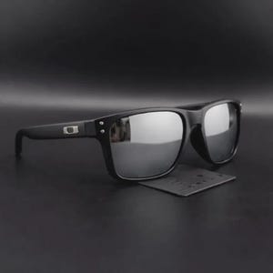 May include: Black sunglasses with mirrored lenses. The sunglasses have a rectangular frame and a small logo on the temple. The lenses reflect the surrounding environment. A small black cleaning cloth is beneath the sunglasses.