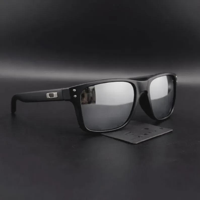 May include: Black sunglasses with mirrored lenses. The sunglasses have a rectangular frame and a small logo on the temple. The lenses reflect the surrounding environment. A small black cleaning cloth is beneath the sunglasses.