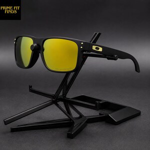 May include: Black sunglasses with gold mirrored lenses are displayed on a black stand. The sunglasses have a square frame with gold accents and the text "POLARIZED" is visible on the lens. The stand has a unique geometric design.