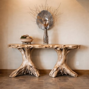 Op de afbeelding: Een lichtbruine houten console met een blad met een natuurlijke rand en twee poten in de vorm van boomstammen. De tafel heeft een rustiek ontwerp. Een decoratieve kom en vaas met takken staan op de tafel. De tafel is ongeveer 180 cm lang.