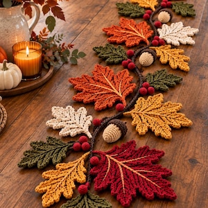 Op de afbeelding: Een lange, decoratieve tafelloper met een gehaakte rank met diverse herfstkleurige bladeren in rood, oranje, geel, crème en groen, geaccentueerd met kleine rode bessen en gehaakte eikeltjes, uitgestald op een houten eettafel.
