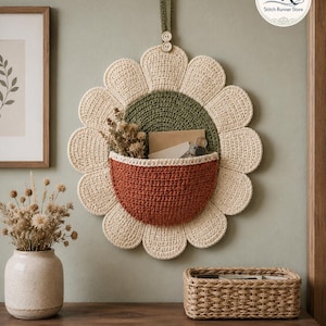 May include: A wall-hanging crochet flower organizer featuring cream-colored petals, a dark green center, and a rust-colored pocket. The piece includes a green hanging loop with two small buttons and contains dried flowers, a note, and metal keys.
